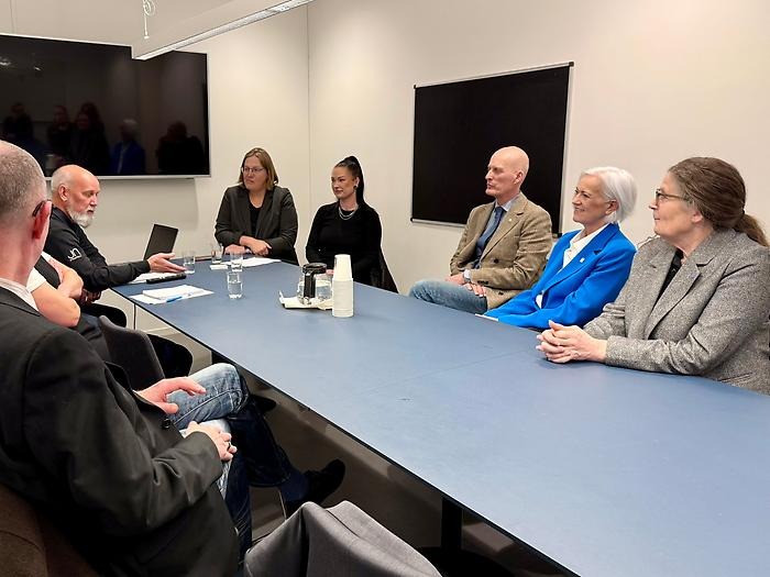 Tiotal människor runt konferensbord, tittar intresserat på talaren längst fram