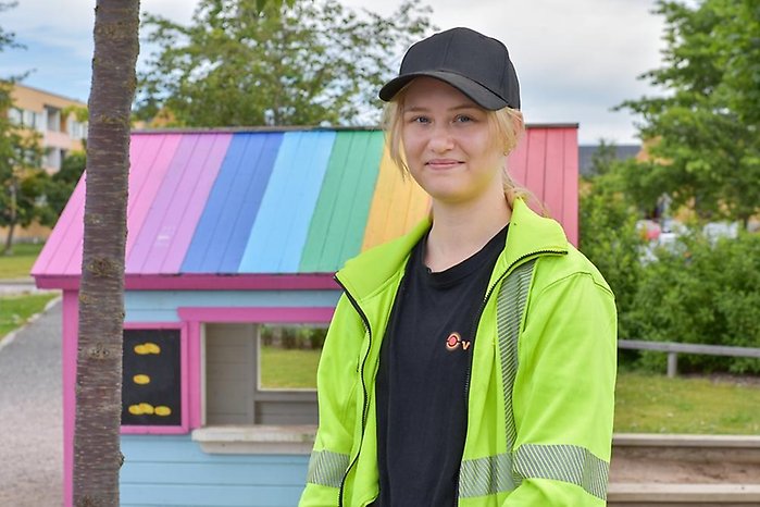 Porträtt av person i svart keps och gul varseljacka framför liten lekstuga med regnbågstak.