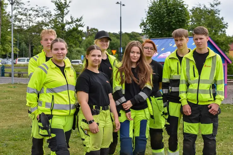 Grupp ungdomar i gula varselkläder står utomhus vid lekplats, träd och parkering i bakgrunden.