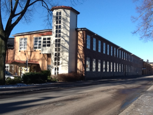 Byggnad på två våningar med tegel och ljusa fasader, träd längs gatan, soligt och lugnt område.