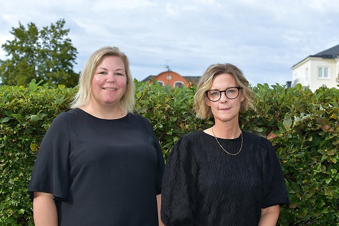 Två personer står framför en grön häck med träd och byggnader i bakgrunden. Båda bär svarta överdelar, och en av dem har glasögon. Himlen är delvis molnig.