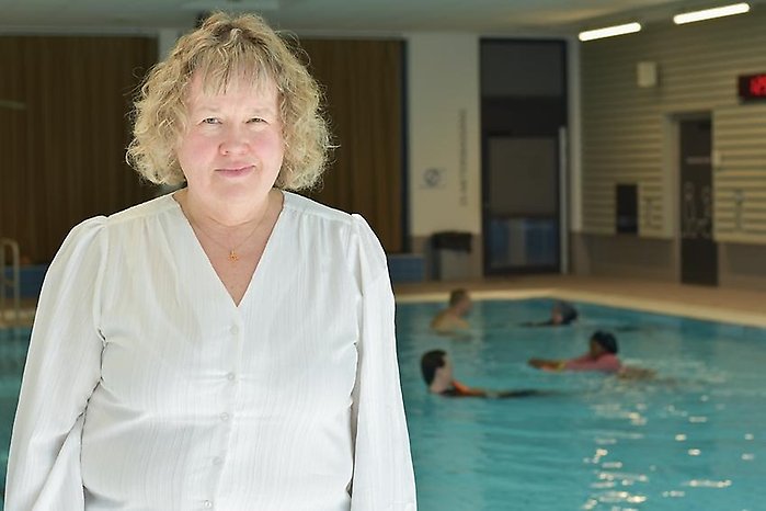 Kvinna vid en inomhusbassäng, med flera personer som simmar i bakgrunden.